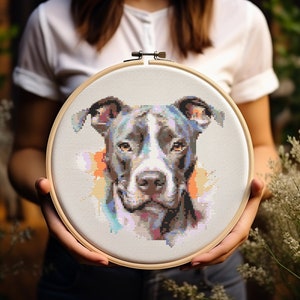 Puede incluir: Un aro de bordado de punto de cruz con un retrato colorido de un perro pitbull. El pelaje del perro es una mezcla de negro, blanco y gris, y el fondo es una acuarela de color rosa, azul y naranja.