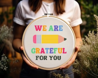 Op de afbeelding: Een borduurring met de tekst "WE ARE GRATEFUL FOR YOU" en een potloodontwerp. De tekst en het potlood zijn in heldere kleuren op een witte achtergrond, geplaatst in een houten ring.
