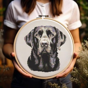 Pode incluir: Um padrão de ponto cruz de um Labrador Retriever preto em um aro de bordado de madeira. O cão está olhando diretamente para o espectador com uma expressão séria.