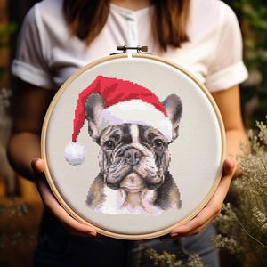 May include: Cross-stitch embroidery of a French bulldog wearing a red Santa hat. The dog is stitched in shades of brown, black, and white. The embroidery is framed in a wooden hoop.