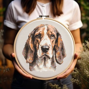May include: A cross-stitch embroidery of a beagle dog's face, with brown, white, and black pixelated details. The artwork is stretched within a round wooden embroidery hoop, held against a blurred natural background.