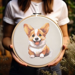 Puede incluir: Un aro de bordado de punto de cruz con una imagen colorida de un perro corgi. El perro sonríe y tiene pelo marrón, blanco y naranja.