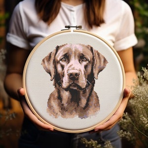 May include: A cross-stitch embroidery of a brown Labrador Retriever dog in a wooden hoop. The dog's portrait is detailed with shades of brown and tan, showcasing its expressive eyes and fur. The hoop is held against a blurred green background.