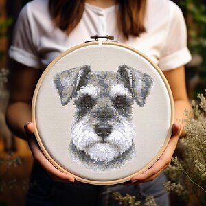 Op de afbeelding: Een ronde houten borduurring met een kruissteekportret van een Schnauzer. De hond is afgebeeld in grijstinten, zwart en wit, met zijn kenmerkende borstelige wenkbrauwen en baard, gecentreerd op een lichtbeige stof.