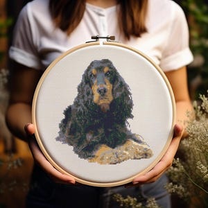 Puede incluir: Un aro de bordado de punto de cruz con un perro cocker spaniel negro y marrón. El perro está sentado con la cabeza inclinada y mirando al espectador.