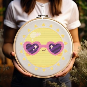 May include: A white embroidery hoop with a yellow sun wearing pink heart-shaped sunglasses with purple lenses. The sun has a happy face and is surrounded by yellow rays.