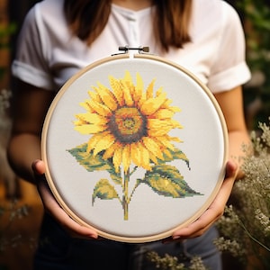 Może przedstawiać: Zbliżenie na gotowy projekt haftu krzyżykowego w drewnianym obręczy. Wzór przedstawia żółty słonecznik z zielonymi liśćmi na białym tle.