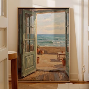 May include: A framed rectangular wall art print depicting an open green door leading to a sunlit terrace overlooking the ocean. The scene features a wooden floor, terracotta pots, a small wooden table, and a view of blue sea waves under a sky.