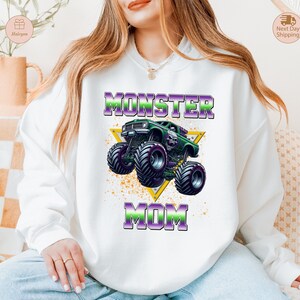 May include: White sweatshirt featuring a graphic of a green monster truck with purple and green text that reads "MONSTER MOM". The design includes a yellow triangle and a splatter effect.