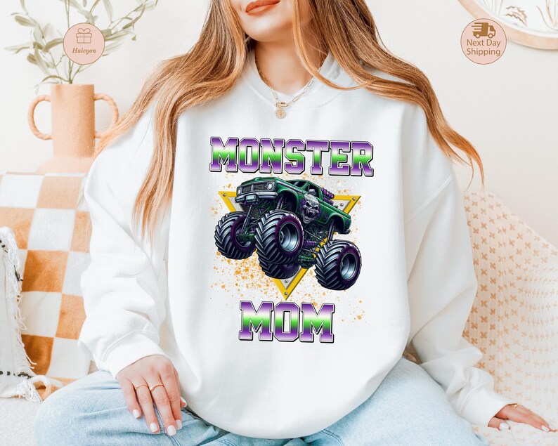 May include: White sweatshirt featuring a graphic of a green monster truck with purple and green text that reads "MONSTER MOM". The design includes a yellow triangle and a splatter effect.