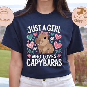 May include: Navy blue t-shirt with the text "JUST A GIRL WHO LOVES CAPYBARAS". The shirt features a cartoon capybara with a pink bow, surrounded by hearts and flowers in pink, teal, and white.