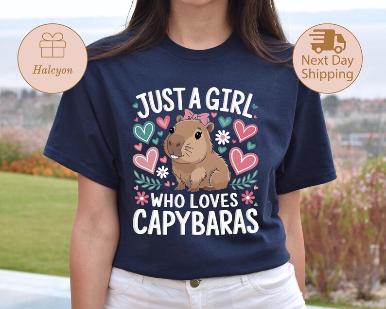 May include: Navy blue t-shirt with the text "JUST A GIRL WHO LOVES CAPYBARAS". The shirt features a cartoon capybara with a pink bow, surrounded by hearts and flowers in pink, teal, and white.