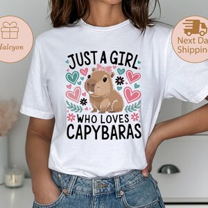 May include: White t-shirt with the text "JUST A GIRL WHO LOVES CAPYBARAS" and a cartoon capybara illustration. The design includes hearts and floral accents in shades of pink, teal, and black. The shirt is a casual wear item.