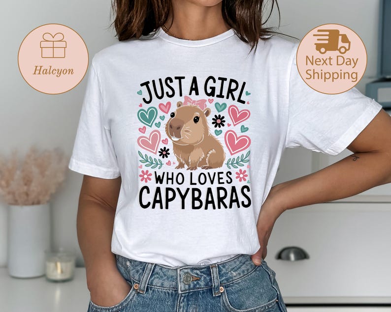May include: White t-shirt with the text "JUST A GIRL WHO LOVES CAPYBARAS" and a cartoon capybara illustration. The design includes hearts and floral accents in shades of pink, teal, and black. The shirt is a casual wear item.