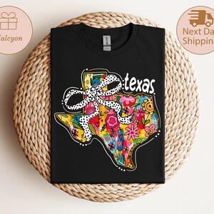 以下が含まれることがあります： カラフルなテキサス州のアウトラインが特徴の黒いTシャツ。花柄と水玉模様のリボンが描かれています。「texas」の文字が白で書かれています。Tシャツは織り込まれた表面に表示されています。