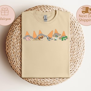 May include: A light tan t-shirt featuring a cartoon design of various animals wearing orange traffic cones. The animals include a pig, a dachshund, a dog, a blue creature, and a green lizard. The shirt is displayed on a woven, circular surface.