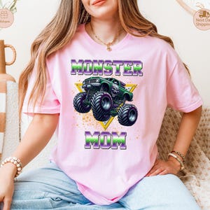 May include: A light pink t-shirt featuring a graphic of a green monster truck inside a yellow triangle. The text "MONSTER MOM" is printed above and below the truck in purple and green. The shirt is worn by a person.