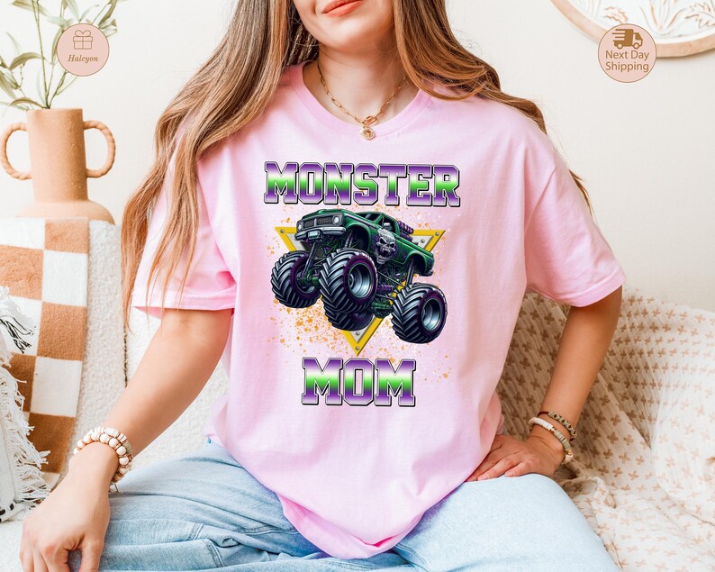 May include: A light pink t-shirt featuring a graphic of a green monster truck inside a yellow triangle. The text "MONSTER MOM" is printed above and below the truck in purple and green. The shirt is worn by a person.