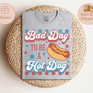 May include: A heather gray t-shirt with the phrase "Bad Day To Be A Hot Dog" in red, white, and blue lettering. A cartoon hot dog graphic is centered below the text. The shirt is displayed on a woven, circular surface.