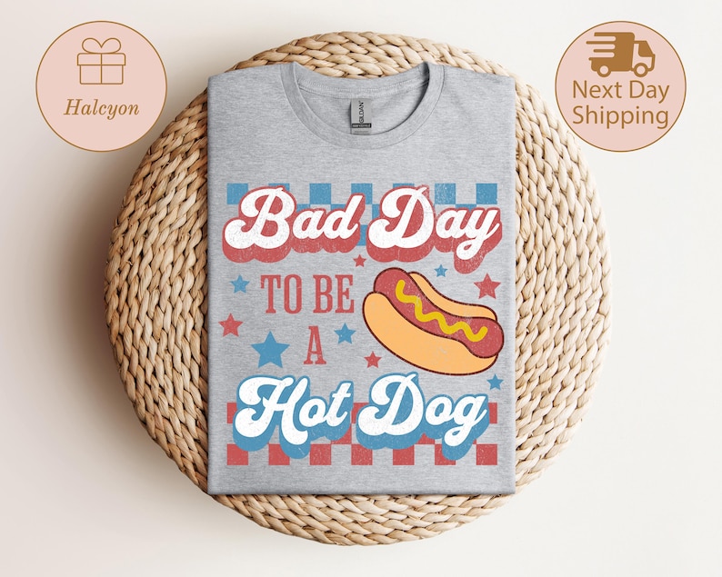May include: A heather gray t-shirt with the phrase "Bad Day To Be A Hot Dog" in red, white, and blue lettering. A cartoon hot dog graphic is centered below the text. The shirt is displayed on a woven, circular surface.
