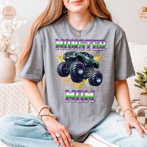 May include: Gray t-shirt featuring a graphic of a green monster truck with purple and green accents, and the words "MONSTER MOM" in a matching color scheme. The design includes a yellow triangle and a splatter effect.
