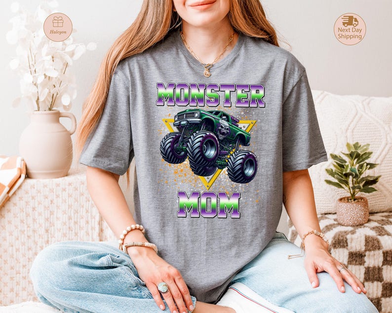 May include: Gray t-shirt featuring a graphic of a green monster truck with purple and green accents, and the words "MONSTER MOM" in a matching color scheme. The design includes a yellow triangle and a splatter effect.
