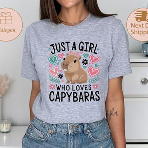 May include: A light gray t-shirt featuring the text "JUST A GIRL WHO LOVES CAPYBARAS" with a cartoon capybara illustration. The design includes hearts and floral accents in pink, green, and blue.