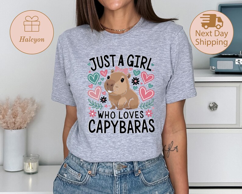 May include: A light gray t-shirt featuring the text "JUST A GIRL WHO LOVES CAPYBARAS" with a cartoon capybara illustration. The design includes hearts and floral accents in pink, green, and blue.