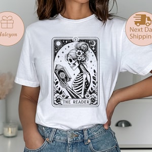 May include: White t-shirt featuring a black and white illustration of a skeleton woman reading a book. The illustration is in the style of a tarot card and says "The Reader" at the bottom.