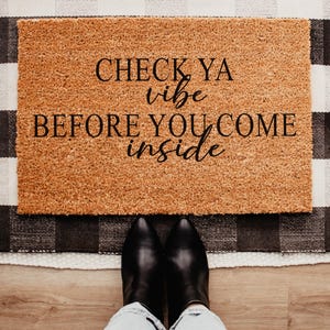 May include: A rectangular tan coir doormat with black text that reads CHECK YA vibe BEFORE YOU COME inside. The mat has a coarse, fibrous texture and is positioned on a black and white buffalo check rug on a light wood floor.
