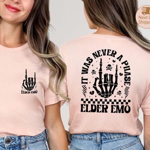 May include: A light pink t-shirt with a black skeleton hand graphic making a rock and roll hand gesture. The text "ELDER EMO" is below the graphic. The back of the shirt has a larger graphic with the text "IT WAS NEVER A PHASE" and "ELDER EMO".