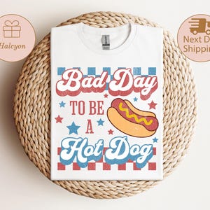 May include: White t-shirt with the text "Bad Day To Be A Hot Dog" in red and blue retro font, with a hot dog graphic. The shirt is displayed on a woven surface.