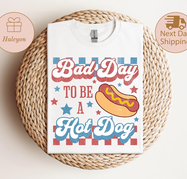 May include: White t-shirt with the text "Bad Day To Be A Hot Dog" in red and blue retro font, with a hot dog graphic. The shirt is displayed on a woven surface.