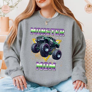 May include: A gray sweatshirt with a graphic of a green monster truck with purple and green accents. The text "MONSTER MOM" is above and below the truck in a purple and green gradient.