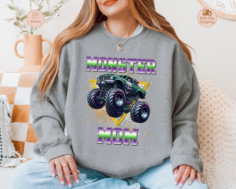 May include: A gray sweatshirt with a graphic of a green monster truck with purple and green accents. The text "MONSTER MOM" is above and below the truck in a purple and green gradient.
