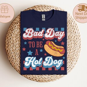 May include: Navy blue t-shirt with the text "Bad Day To Be A Hot Dog" in red, white, and blue. The design includes a cartoon hot dog and stars. The shirt is displayed on a woven surface.