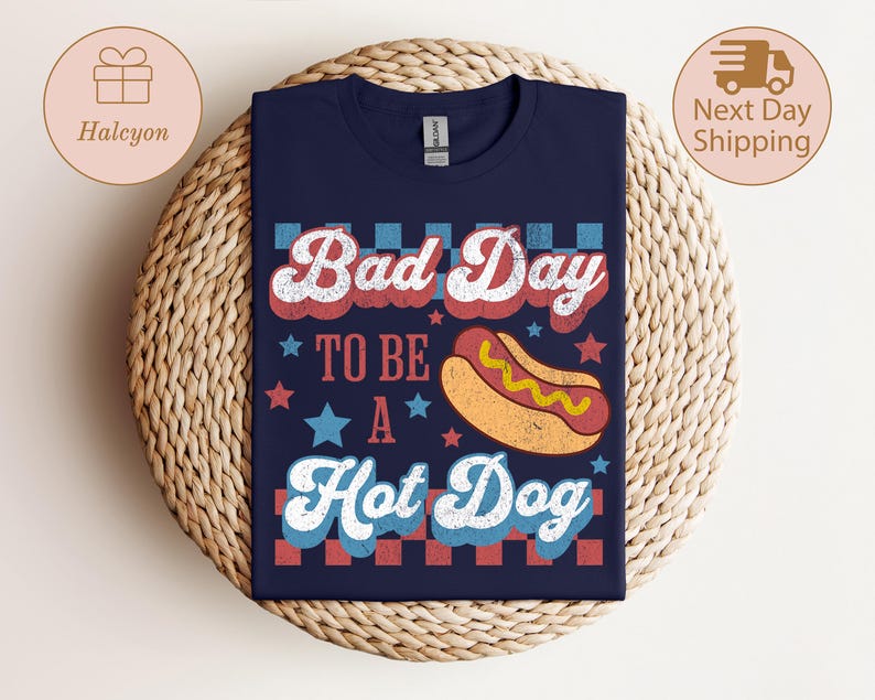 May include: Navy blue t-shirt with the text "Bad Day To Be A Hot Dog" in red, white, and blue. The design includes a cartoon hot dog and stars. The shirt is displayed on a woven surface.
