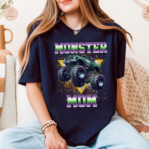 May include: Navy blue t-shirt with a graphic of a green monster truck and the words "MONSTER MOM" in purple and green. The design includes a yellow triangle and a splatter effect.