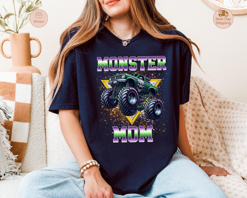 May include: Navy blue t-shirt with a graphic of a green monster truck and the words "MONSTER MOM" in purple and green. The design includes a yellow triangle and a splatter effect.