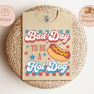 May include: Beige t-shirt with the text "Bad Day To Be A Hot Dog" in red, white, and blue. A cartoon hot dog graphic is featured below the text. The shirt has a retro, vintage design.
