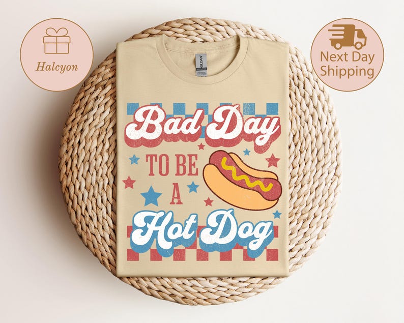 May include: Beige t-shirt with the text "Bad Day To Be A Hot Dog" in red, white, and blue. A cartoon hot dog graphic is featured below the text. The shirt has a retro, vintage design.