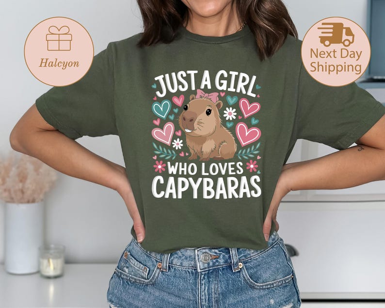 May include: Olive green t-shirt with the text "JUST A GIRL WHO LOVES CAPYBARAS" in white, pink, and blue. The design includes a cartoon capybara, hearts, and floral accents. The shirt is a comfortable, casual style.