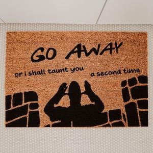 May include: A rectangular doormat with a brown, textured surface. The mat features the words "GO AWAY" in large black letters, with the phrase "or I shall taunt you a second time" below. A black silhouette of a person is in the center.
