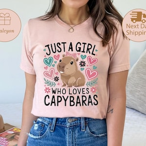 May include: A light pink t-shirt with the text "JUST A GIRL WHO LOVES CAPYBARAS" in black. The design includes a cartoon capybara, hearts, and floral accents. The shirt is made of a soft material.