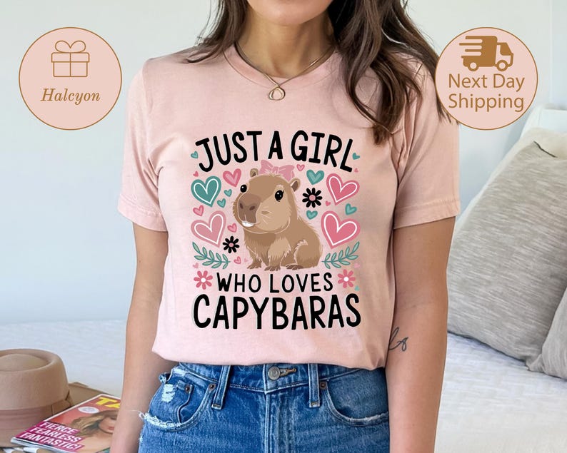 May include: A light pink t-shirt with the text "JUST A GIRL WHO LOVES CAPYBARAS" in black. The design includes a cartoon capybara, hearts, and floral accents. The shirt is made of a soft material.