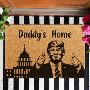 May include: A rectangular doormat with the text "Daddy's Home" and a silhouette of a building and a man giving a thumbs-up. The doormat is made of a natural fiber material and is placed on a black and white striped surface.