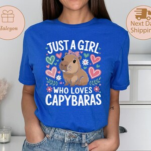 May include: A royal blue t-shirt with the text "JUST A GIRL WHO LOVES CAPYBARAS" in white. The graphic features a cartoon capybara with a pink bow, surrounded by hearts and flowers. The shirt is a crew neck.