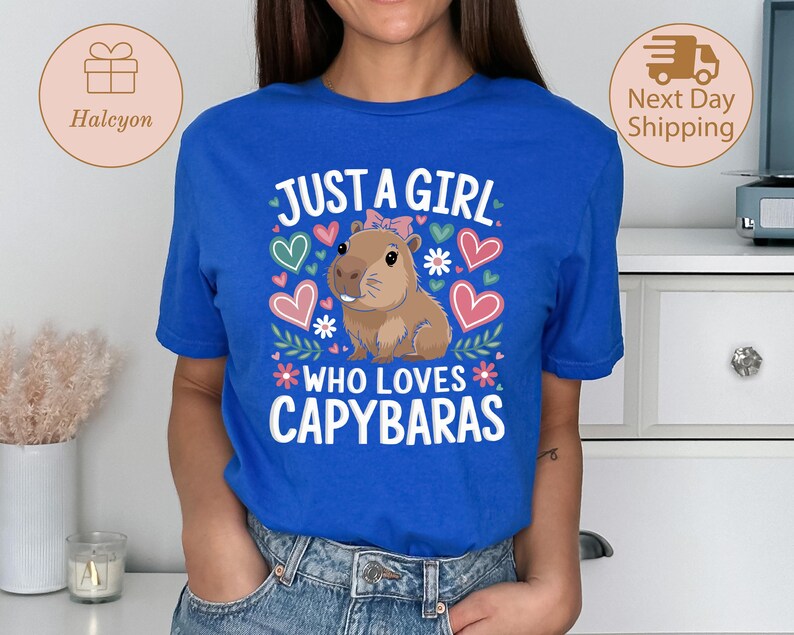 May include: A royal blue t-shirt with the text "JUST A GIRL WHO LOVES CAPYBARAS" in white. The graphic features a cartoon capybara with a pink bow, surrounded by hearts and flowers. The shirt is a crew neck.
