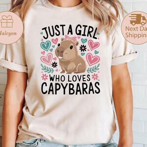 May include: Beige t-shirt featuring a capybara illustration with the text "JUST A GIRL WHO LOVES CAPYBARAS." The design includes pink and teal hearts, flowers, and leaves. The capybara has a pink bow.