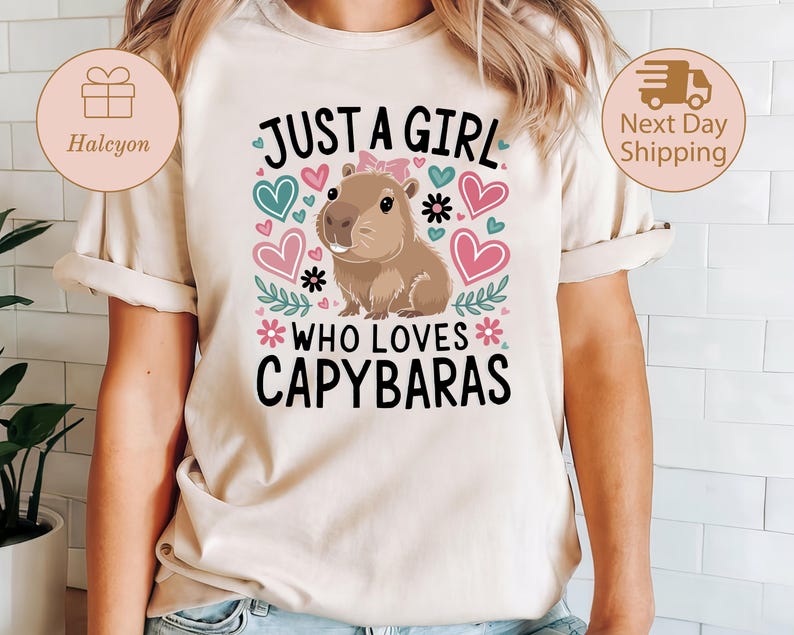 May include: Beige t-shirt featuring a capybara illustration with the text "JUST A GIRL WHO LOVES CAPYBARAS." The design includes pink and teal hearts, flowers, and leaves. The capybara has a pink bow.